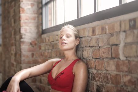 A young woman in activewear rests against a brick wall after exercising, showcasing fitness and dedication.