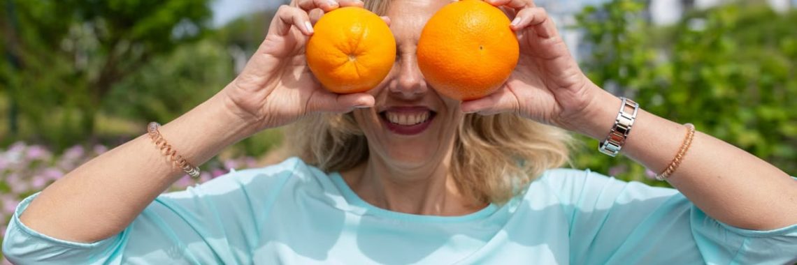 A smiling woman playfully holding oranges in a sunny park setting.